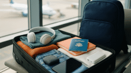 Mala de mão organizada sobre assento de aeroporto, com roupas dobradas, eletrônicos, passaporte e mochila ao fundo