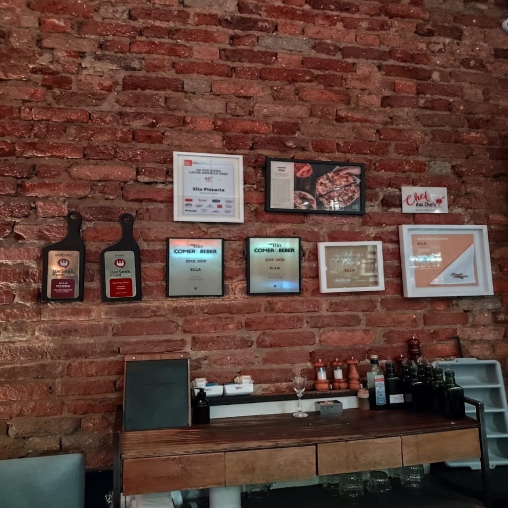 Rustic brick wall at Ella Pizzaria decorated with framed awards and photos, including recognitions like “Top Pizza in Latin America” and “Comer & Beber” honors.