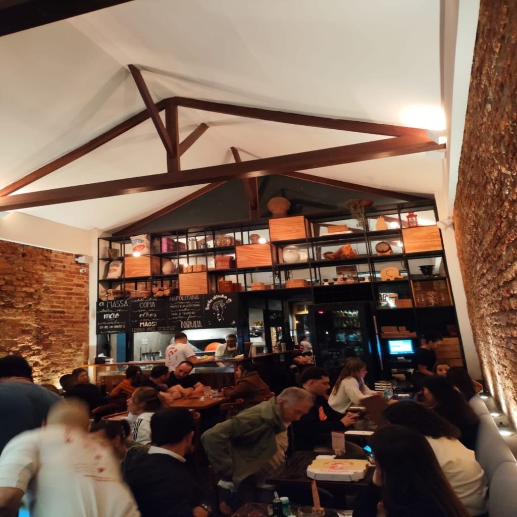 Cozy interior of Ella Pizzaria with a high ceiling, wooden beams, and exposed brick walls, showing a bustling dining area filled with guests enjoying the lively atmosphere.