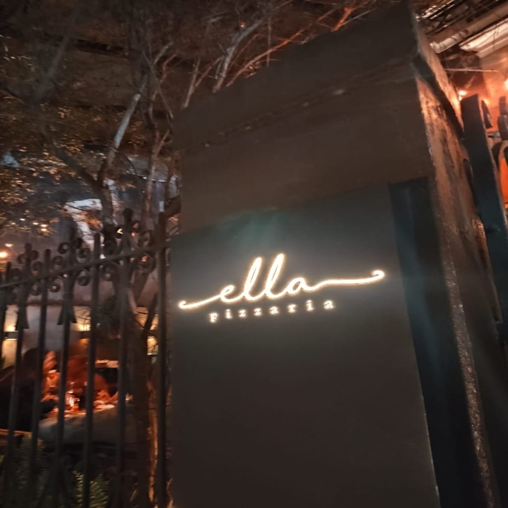 Nighttime view of Ella Pizzaria’s entrance, featuring a glowing white sign and a charming tree-lined exterior in Rio de Janeiro’s Jardim Botânico neighborhood.