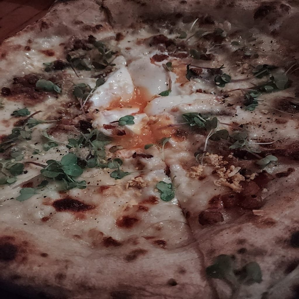 Close-up of an artisanal pizza from Ella Pizzaria featuring a Neapolitan crust, melted cheese, runny egg yolk, and fresh microgreens on top.