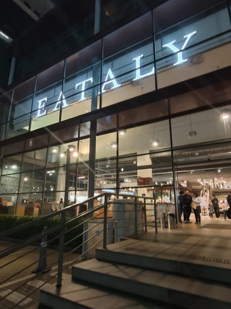 Vista da entrada principal do Eataly S&atilde;o Paulo &agrave; noite com fachada de vidro e letreiro iluminado
