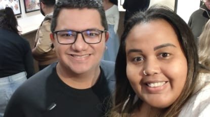 Casal sorridente no foyer do Teatro Clara Nunes, com exposição de cartazes ao fundo e público circulando