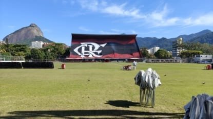 Campo de futebol do Flamengo na Gávea, com arquibancada rubro-negra e o Morro Dois Irmãos ao fundo, em dia ensolarado