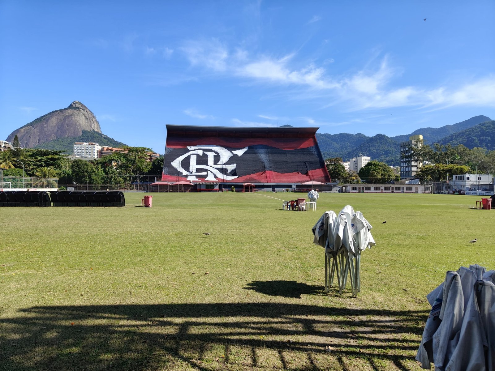 Campo de futebol do Flamengo na Gávea, com arquibancada rubro-negra e o Morro Dois Irmãos ao fundo, em dia ensolarado