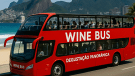 Ônibus vermelho de dois andares chamado Wine Bus estacionado à beira-mar, com turistas participando de uma degustação panorâmica de vinhos, tendo ao fundo as praias e montanhas do Rio de Janeiro, incluindo o Morro Dois Irmãos.