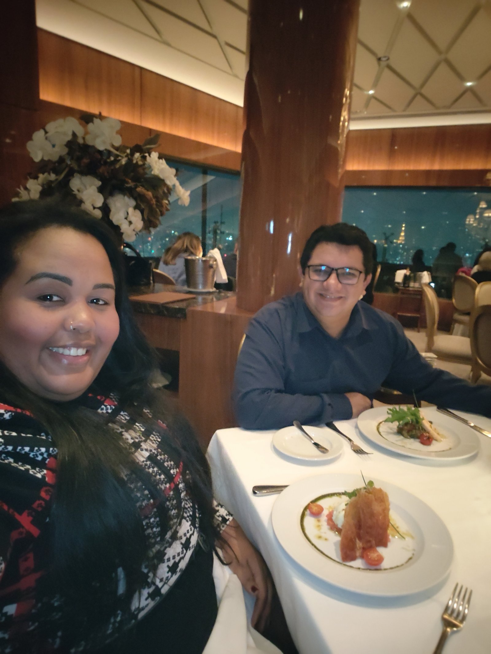 Couple dining at Terraco Italia with gourmet dishes on the table and a panoramic nighttime view of São Paulo in the background.