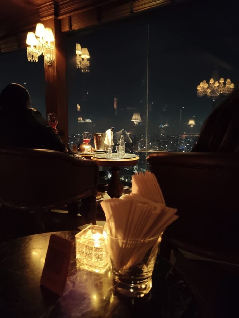 Vista noturna do bar do Terraço Itália com luzes suaves, taças de vinho, velas na mesa e janelas de vidro com São Paulo ao fundo