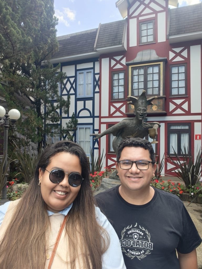 Casal sorrindo em frente à famosa estátua do bobo da corte, símbolo da Vila com fachada colorida ao fundo.