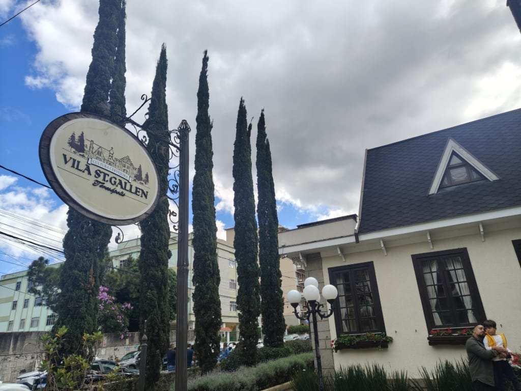 Placa externa da Vila St. Gallen em Teresópolis com pinheiros ao fundo e céu parcialmente nublado, marcando a entrada do complexo de inspiração alemã