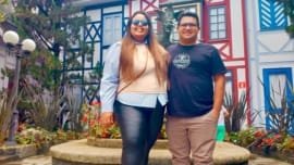 Casal sorridente posando em frente à fonte central da Vila St. Gallen, com fachadas coloridas em estilo bávaro ao fundo.