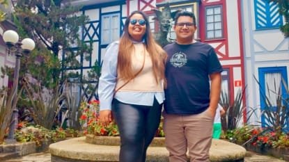 Casal sorridente posando em frente à fonte central da Vila St. Gallen, com fachadas coloridas em estilo bávaro ao fundo.