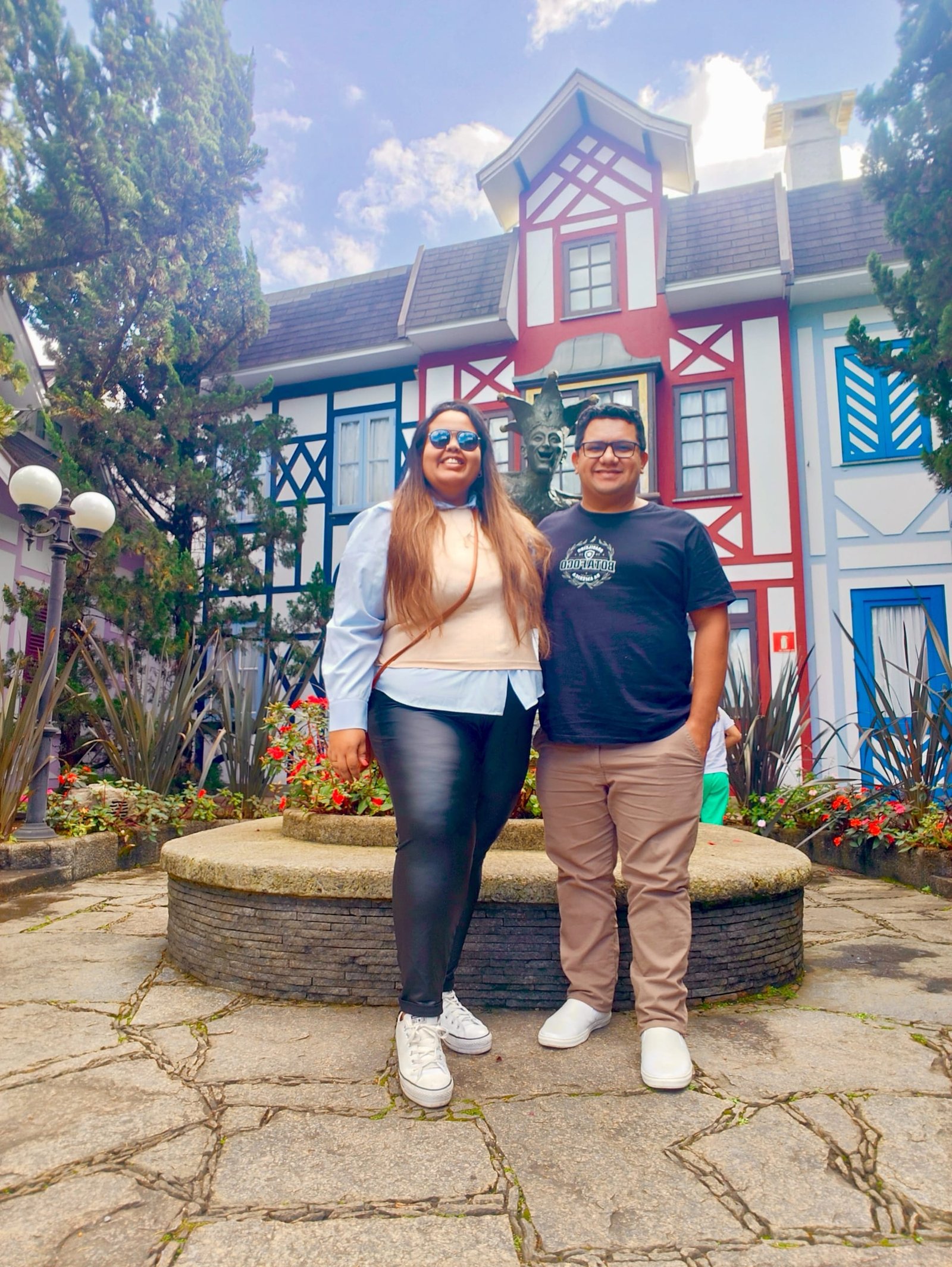 Casal sorridente posando em frente à fonte central da Vila St. Gallen, com fachadas coloridas em estilo bávaro ao fundo.