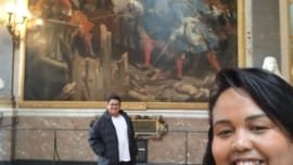 Casal viajando no Palácio de Versalhes durante o Roteiro Europa 2025, sorrindo em frente a uma pintura histórica nas galerias do palácio na França, roteiro de 2 dias em Paris