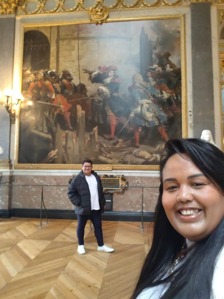 Casal viajando no Palácio de Versalhes durante o Roteiro Europa 2025, sorrindo em frente a uma pintura histórica nas galerias do palácio na França, roteiro de 2 dias em Paris