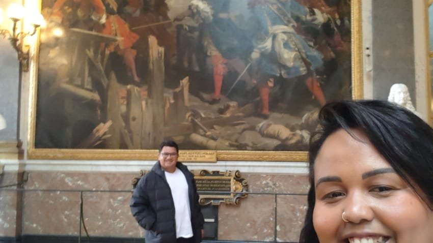 Casal viajando no Palácio de Versalhes durante o Roteiro Europa 2025, sorrindo em frente a uma pintura histórica nas galerias do palácio na França, roteiro de 2 dias em Paris