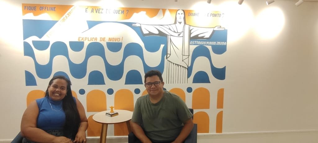 Visitantes sorrindo em frente ao mural colorido, com ilustração do Cristo Redentor e frases divertidas sobre jogos de tabuleiro.