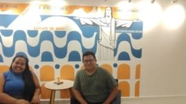 Visitantes sorrindo em frente ao mural colorido da Luderia Carioca, com ilustração do Cristo Redentor e frases divertidas sobre jogos de tabuleiro.