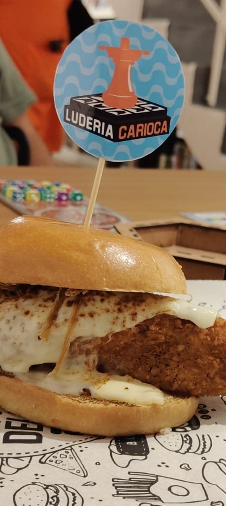 Hambúrguer artesanal servido com bandeirinha personalizada, representando o cardápio saboroso do espaço de jogos de tabuleiro.