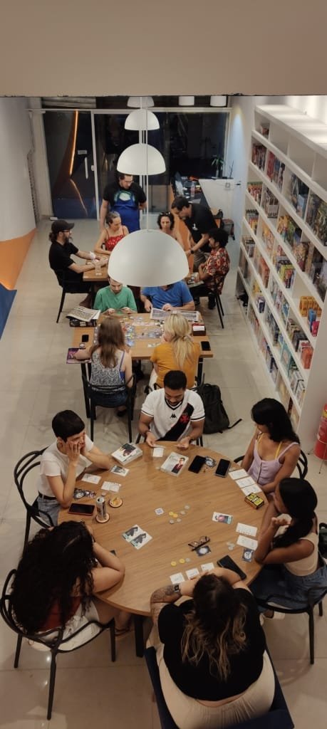 Vista aérea do salão principal da Luderia Carioca, com várias mesas ocupadas por grupos jogando e prateleiras cheias de jogos de tabuleiro.