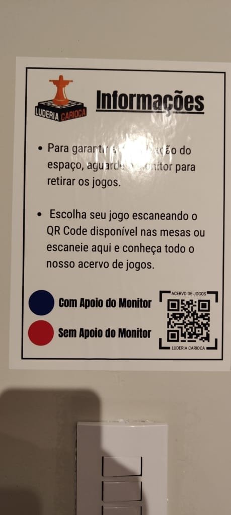 Placa informativa da Luderia Carioca explicando como escolher e retirar jogos de tabuleiro com apoio dos monitores e QR Code de acesso ao acervo.