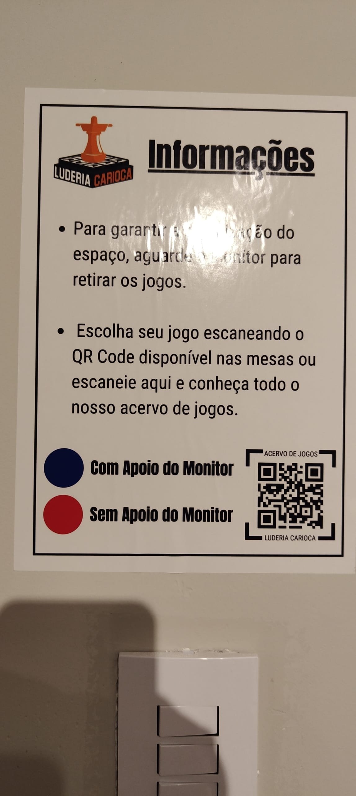 Informational plaque at Luderia Carioca explaining how to choose and take board games with the help of the monitors and the QR Code to access the collection.