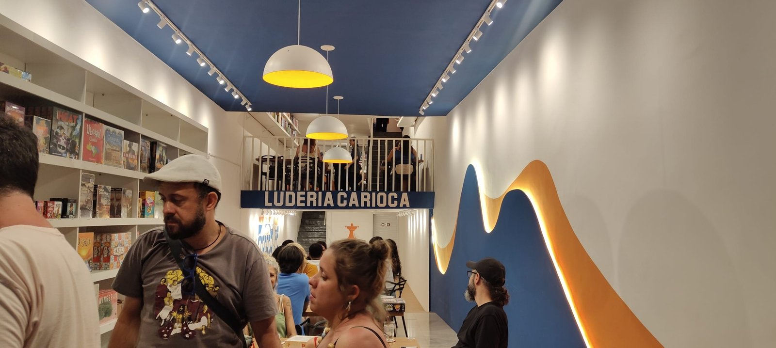 Interior da Luderia Carioca no Rio de Janeiro, com prateleiras cheias de jogos de tabuleiro e visitantes aproveitando o ambiente moderno e acolhedor.