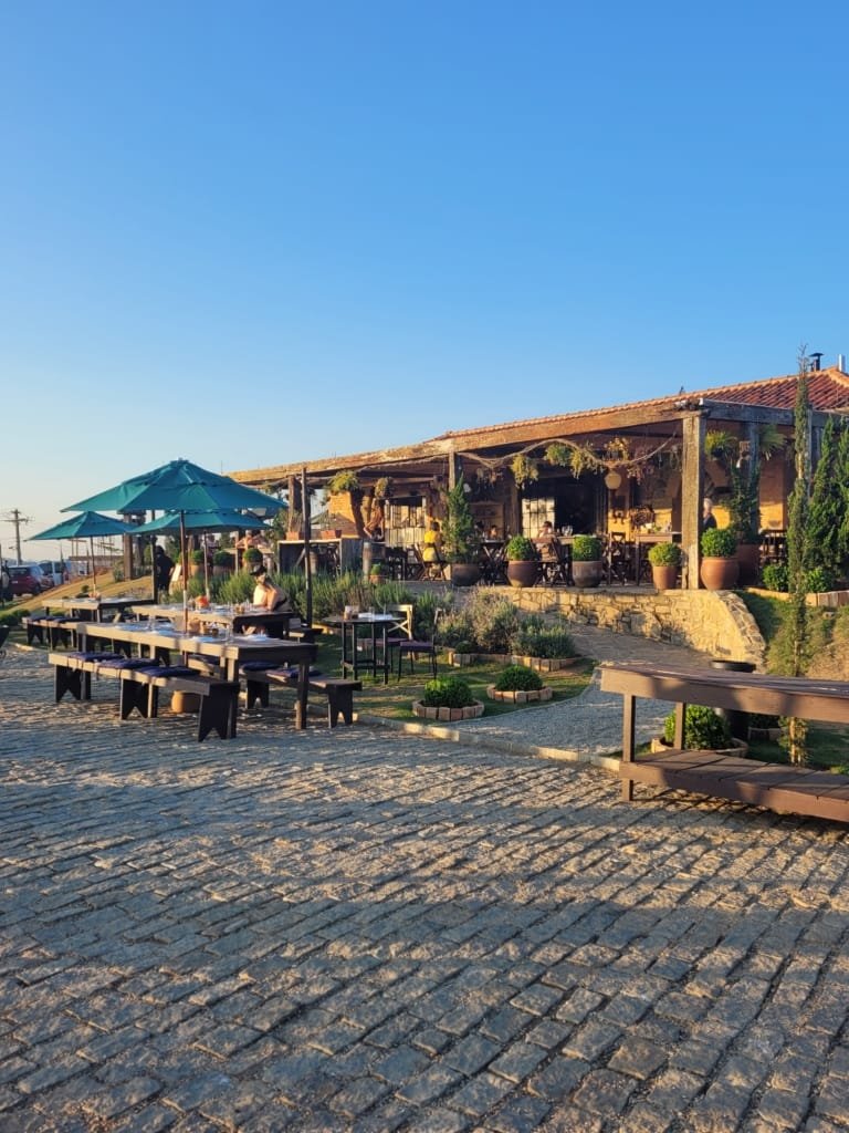 Ambiente externo da Borgo del Vino em Areal com mesas ao ar livre, arquitetura rústica e vista para a serra em um fim de tarde ensolarado