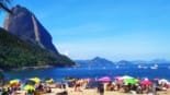 Vista panorâmica de uma das praias do Rio de Janeiro, com pessoas curtindo o dia, mar azul e o Pão de Açúcar ao fundo, um dos principais cartões-postais da cidade.