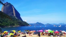Vista panorâmica de uma das praias do Rio de Janeiro, com pessoas curtindo o dia, mar azul e o Pão de Açúcar ao fundo, um dos principais cartões-postais da cidade.