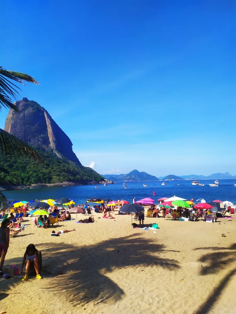 Praias do Rio perto do metrô: guia prático para ir sem carro (2026) 9 Vista panorâmica de uma das praias do Rio de Janeiro, com pessoas curtindo o dia, mar azul e o Pão de Açúcar ao fundo, um dos principais cartões-postais da cidade.