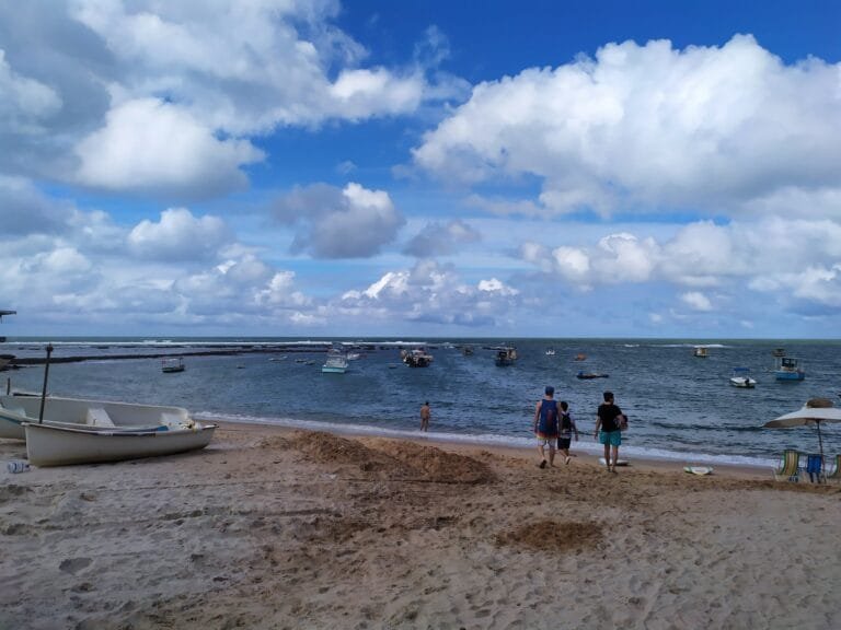 Praia no Nordeste do Brasil com barcos no mar e turistas caminhando na areia